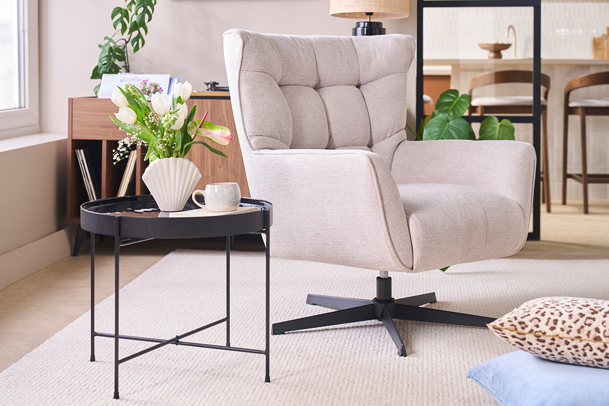 Drehbarer Sessel AVERY aus beigem Stoff, 3/4-Ansicht, mit Tisch und Blumen im Hintergrund.