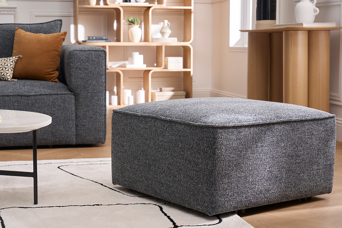 Grauer Stoff-Hocker KERRY als Fu�st�tze, 3/4-Ansicht in einem modernen Wohnzimmer.