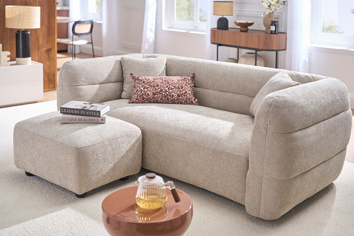 Beiger Stoff-Hocker JOYA als Fu�st�tze, neben einem Sofa, Gesamtansicht in hellem Innenraum.
