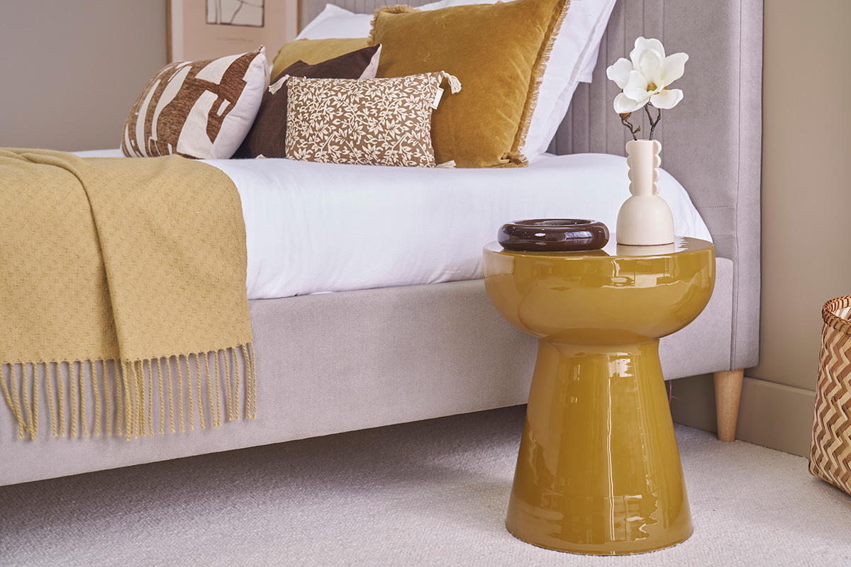 Runder Beistelltisch aus bronzefarbenem emailliertem Metall, neben einem Bett mit Zierkissen.