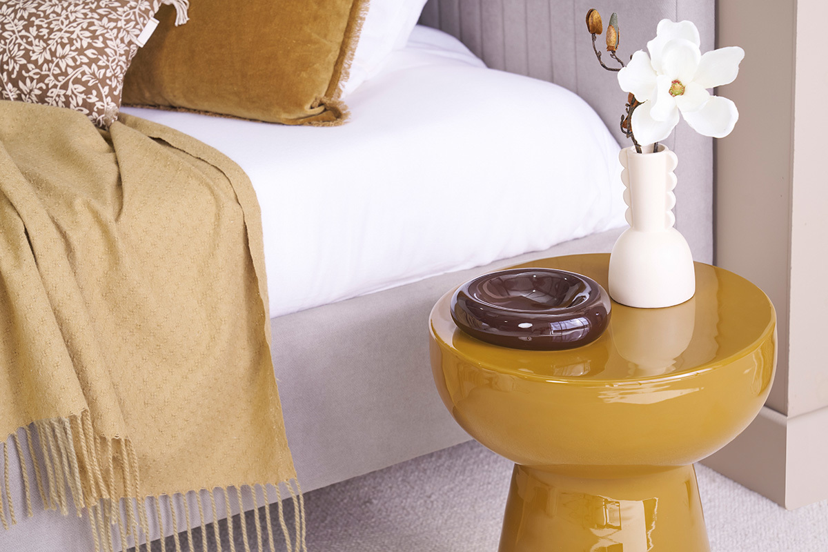 Runder MOGOU Beistelltisch in Bronze mit wei�er Vase und braunem Aschenbecher, neben einem Bett.