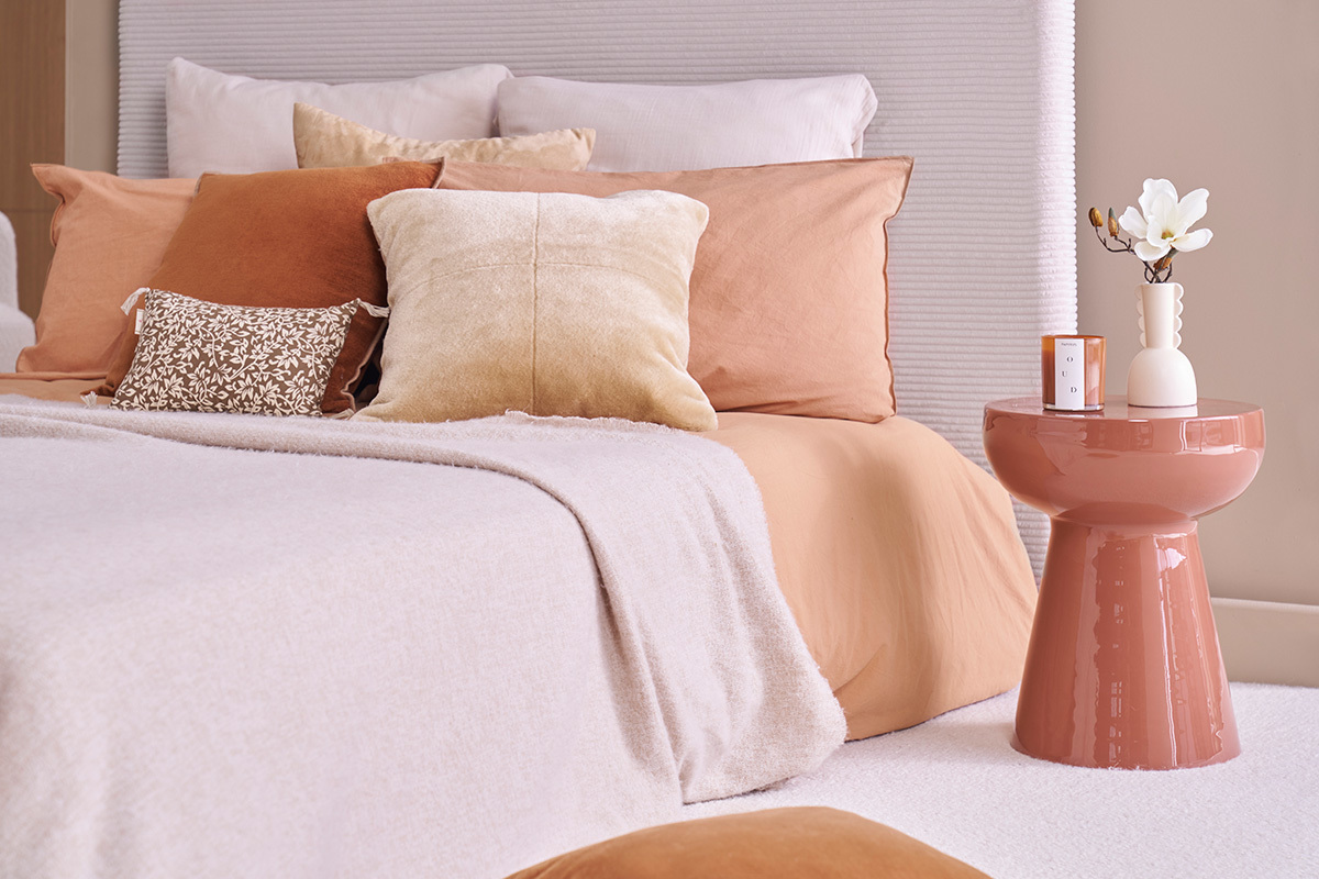 Runder Beistelltisch aus emailliertem Metall in Terrakotta neben einem Bett mit beigen und Terrakotta-Kissen.