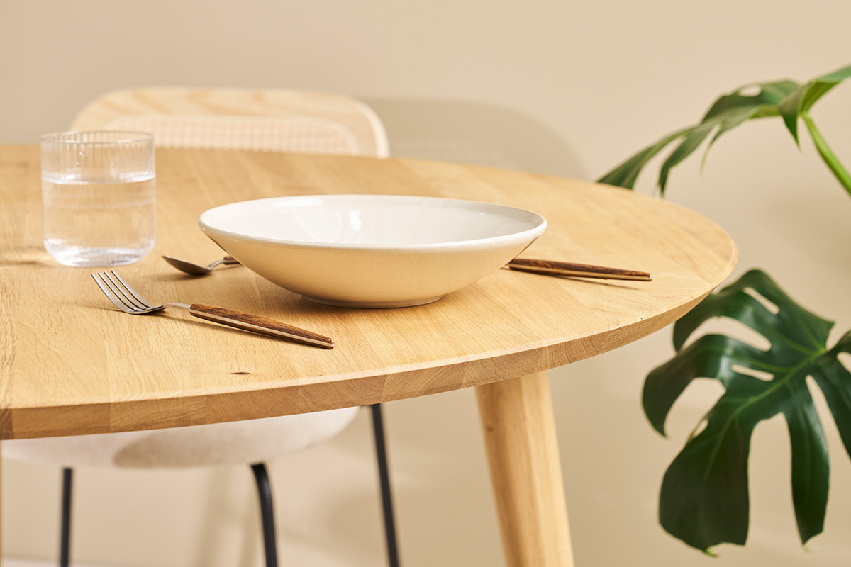 Runder Eichentisch in hellem Holz mit Geschirr und Pflanze im Hintergrund, Nahaufnahme.