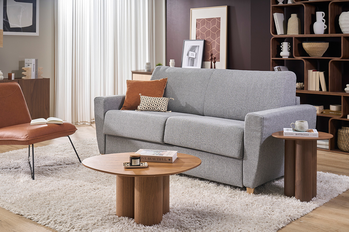 Schlafsofa GRAHAM aus hellgrauem Stoff mit Kissen, begleitet von Couchtischen, Frontalansicht.