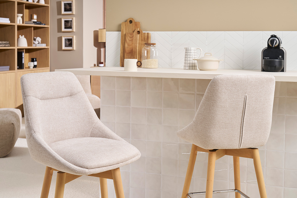 Zwei skandinavische Barhocker aus beigem Stoff und hellem Holz vor einer K�chentheke.