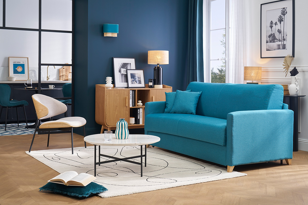 Wohnzimmer mit blauem Schlafsofa, frontal Ansicht, runder Couchtisch und Sessel aus hellem Holz.