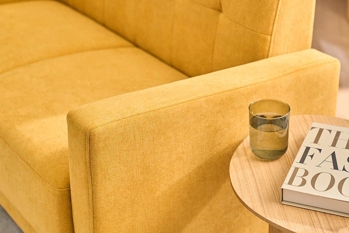 Zweiersofa in Senfgelb mit Samteffekt, Nahaufnahme der Armlehne, Beistelltisch mit Glas und Buch.