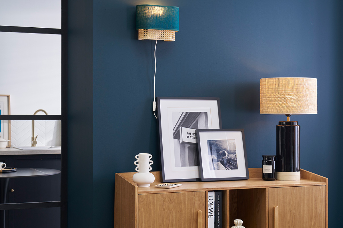 Wandleuchte in Entenblau und Rattan an blauer Wand, neben Bilderrahmen und Lampe auf einem M�belst�ck.